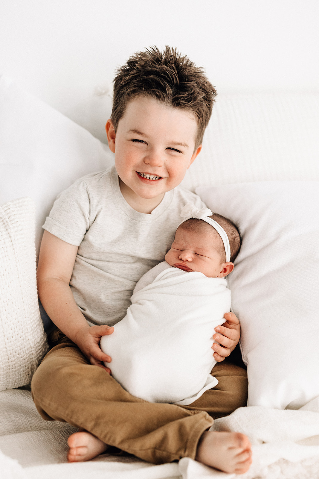 A happy toddler boy sits with his newborn baby sister sleeping in his lap in a white swaddle and bow