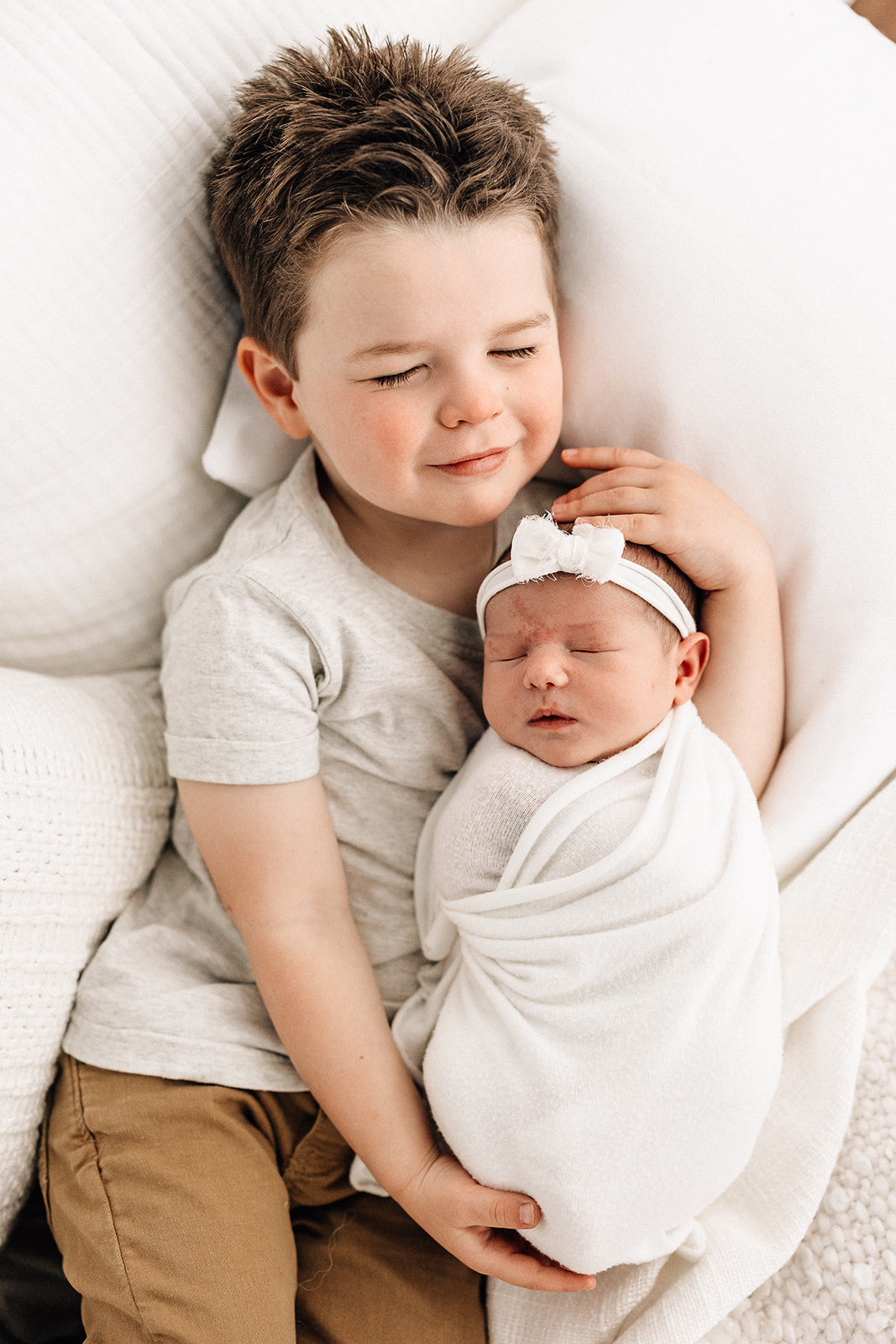 A toddler boy in a grey shirt and brown pants sleeps on a couch while holding his sleeping newborn baby sister in a white swaddle before some Toddler Activities St. Louis