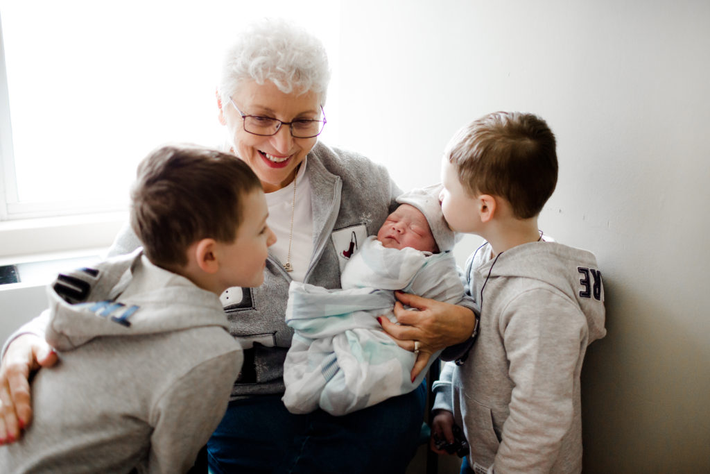Grandma holding newborn baby in hospital. Older sibling kissing newborn on head