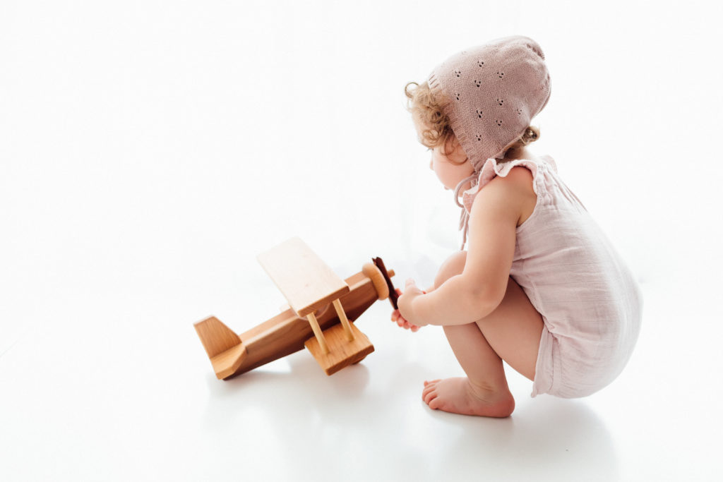Baby in light purple Jamie Kay onesie and bonnet playing with wooden airplane. 