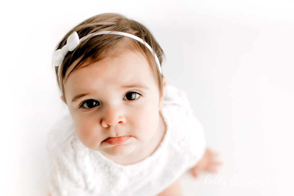 Photo by St Louis Baby Photographer of baby wearing white lace romper and white headband. Looking at camera with pouty lip. 
