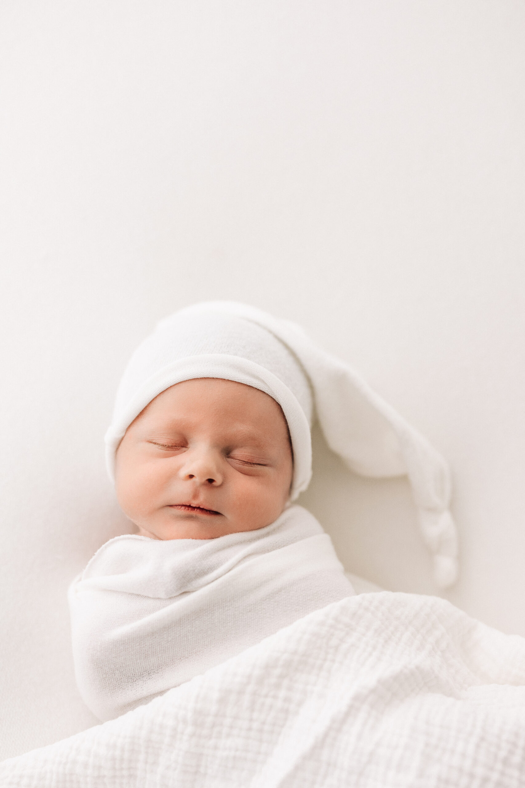 Newborn Baby Boy sleeps while swaddled in white wearing a white sleepy cap