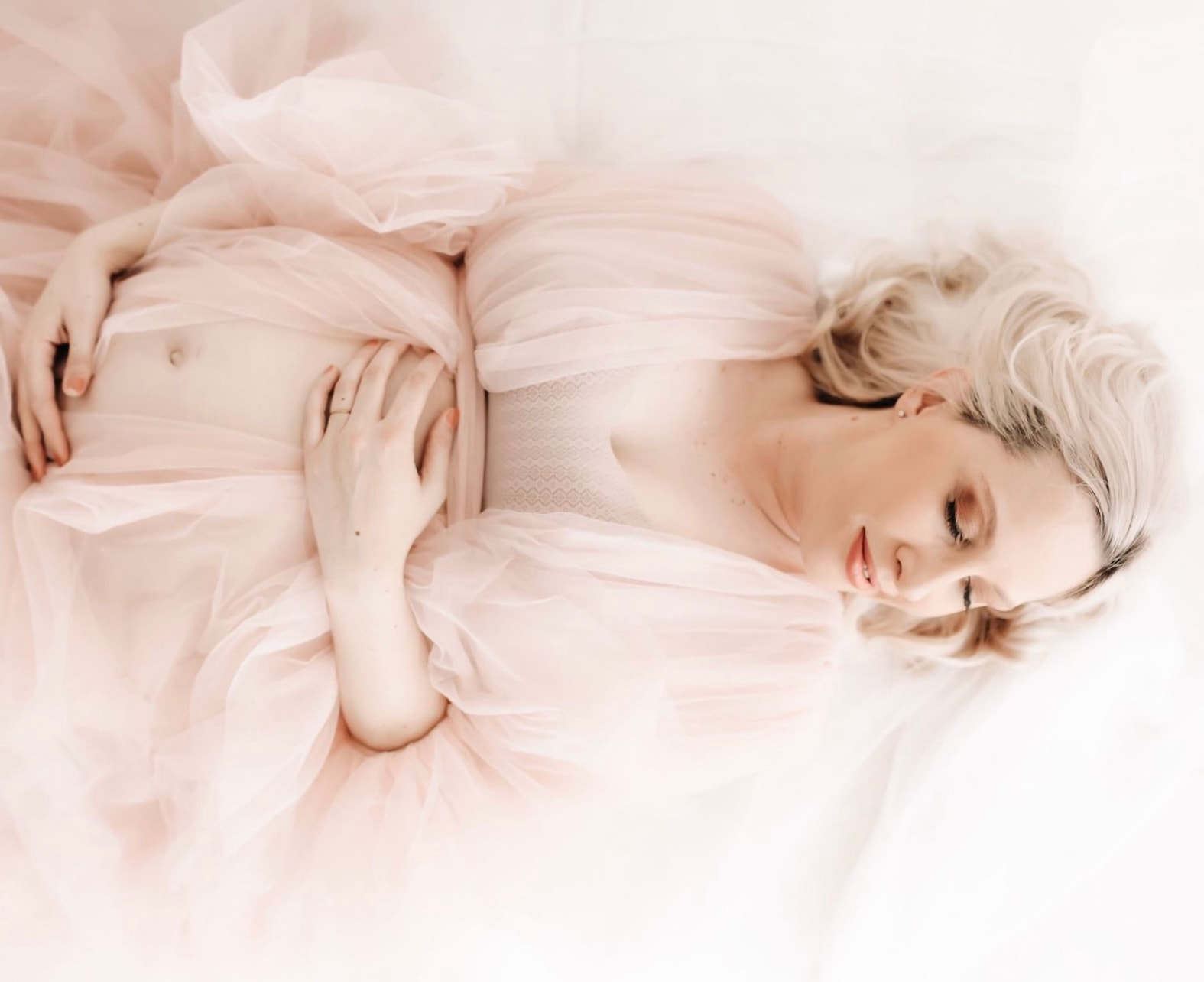 mother to be wearing a pink velour maternity gown, lays on a white bed in a studio