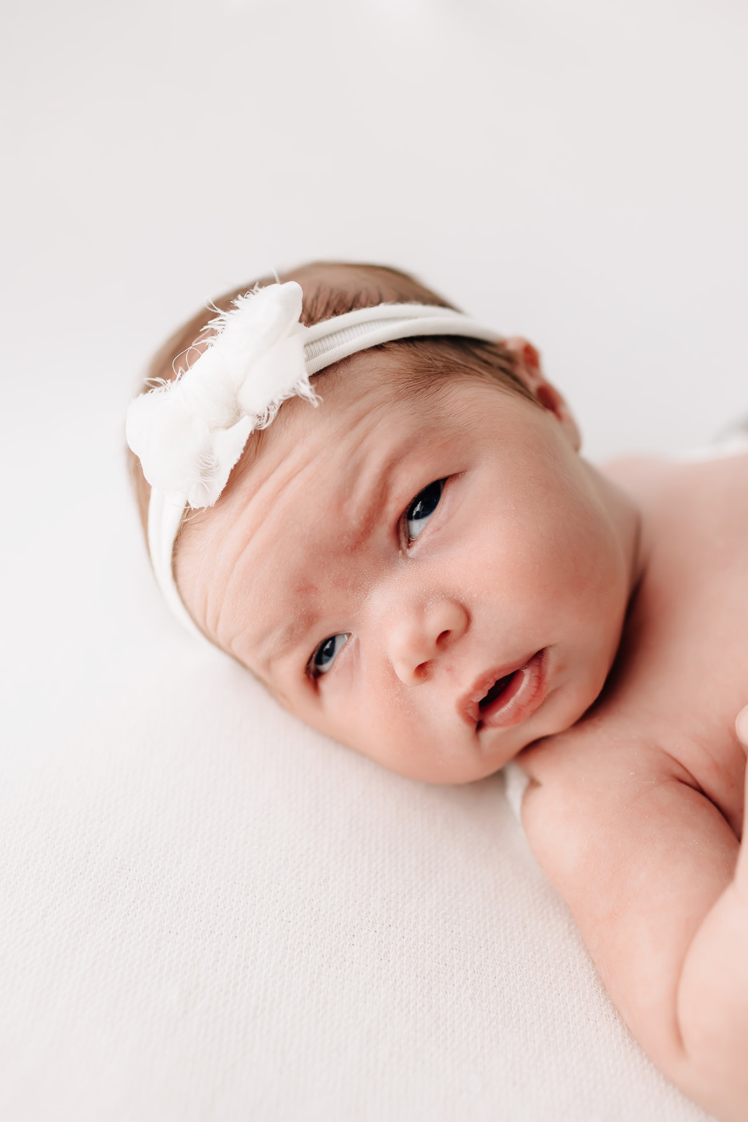 newborn baby lays on a white bed in a white headband with eyes open dancing for birth