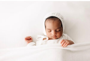 newborn baby sleeps under a white blanket in a white knit bonnet peaceful beginnings midwifery