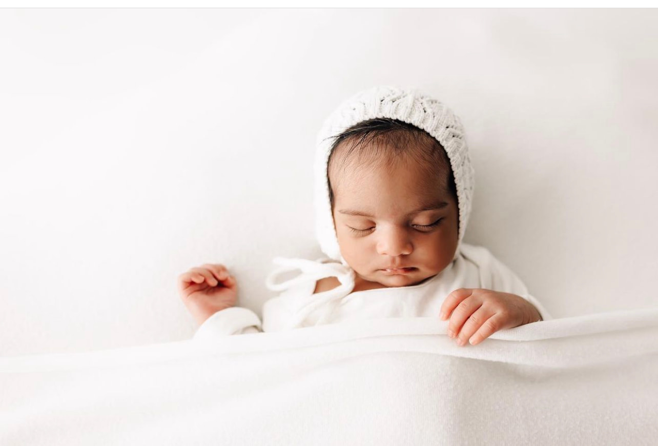 newborn baby sleeps under a white blanket in a white knit bonnet peaceful beginnings midwifery