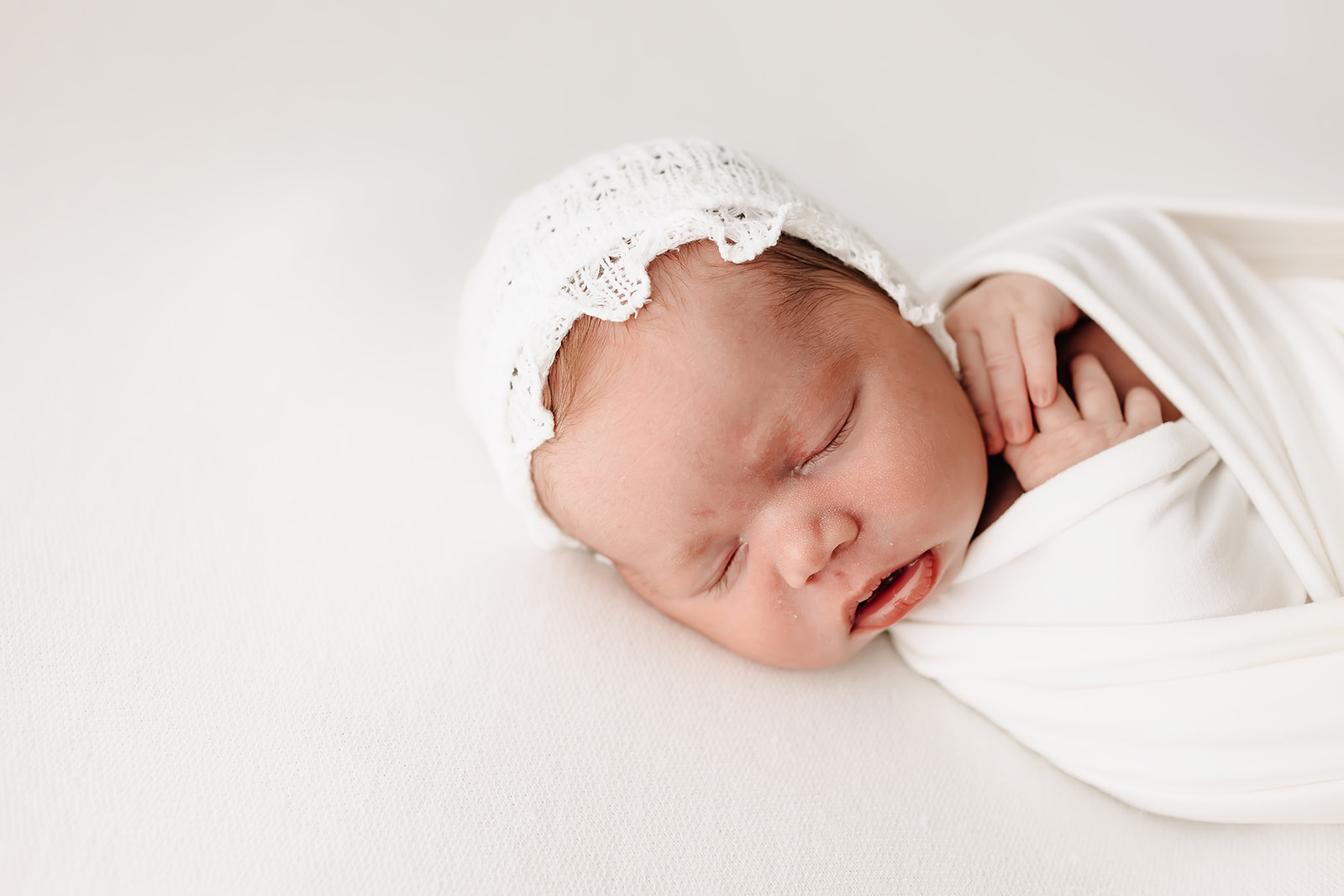 A newborn baby sleeps in a studio on a white bed with hands swaddled under her chin