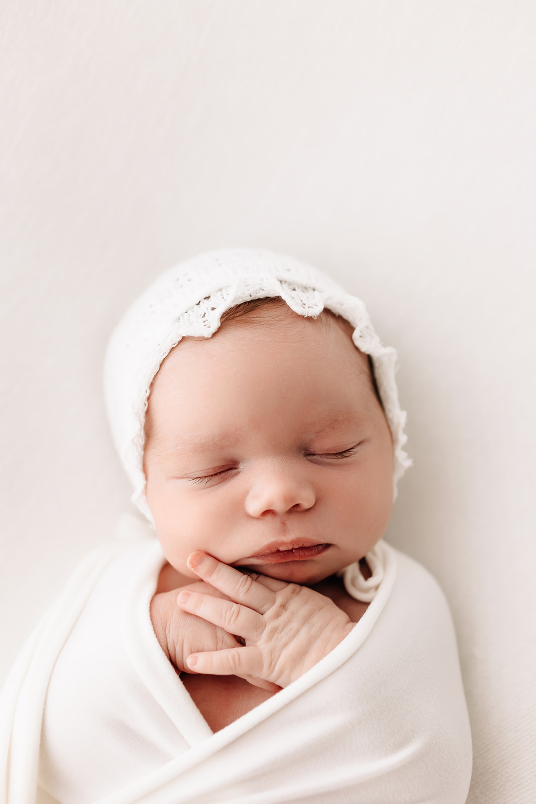 A newborn baby sleeps in a white swaddle and lace bonnet in a studio Washington University OBGYN