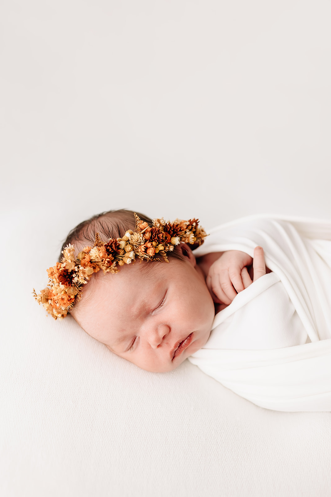A newborn baby sleeps in a white swaddle and floral headband with hands poking out