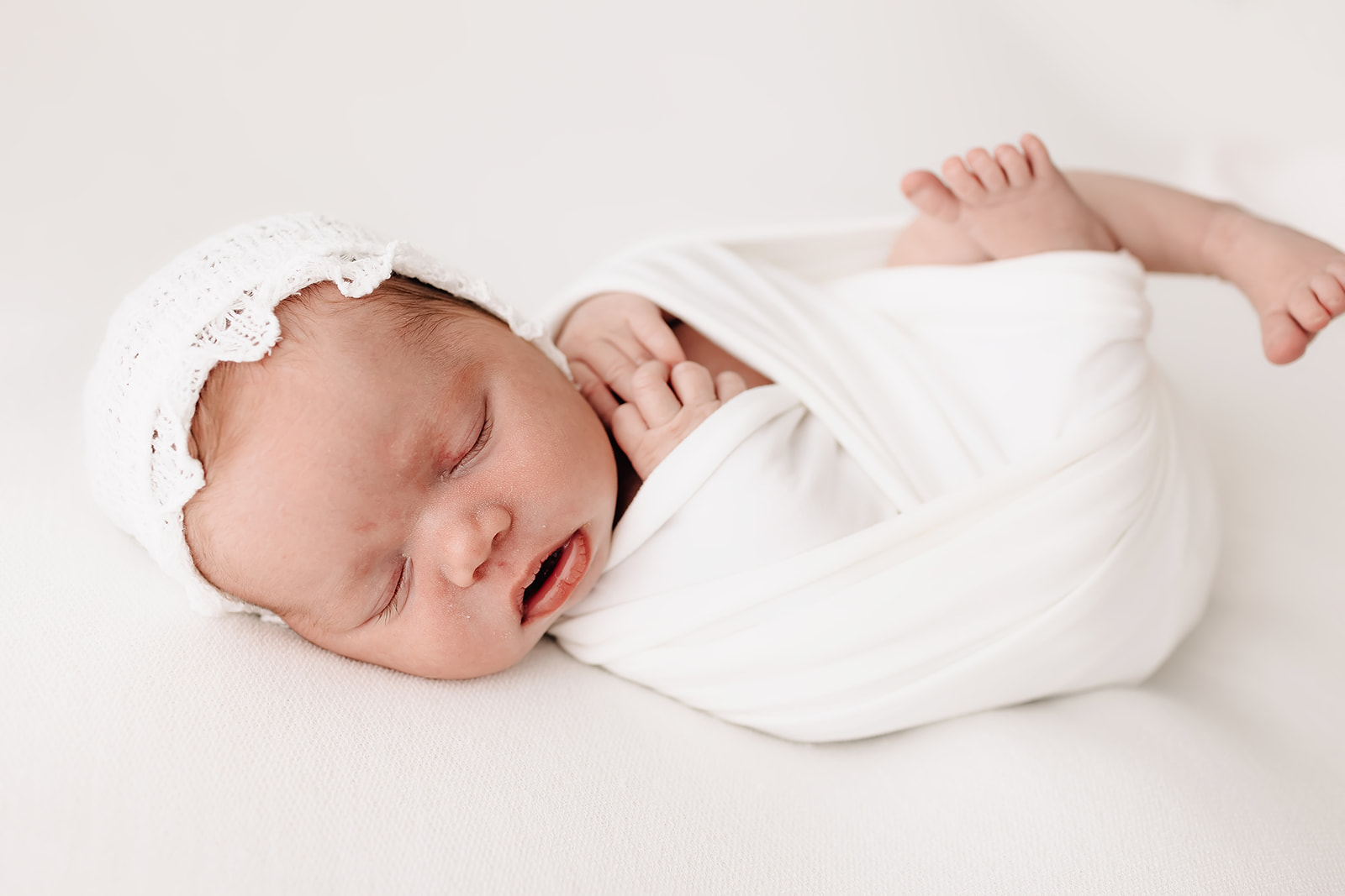 A newborn baby sleeps wrapped up in a white swaddle and lace bonnet Happy Up Inc