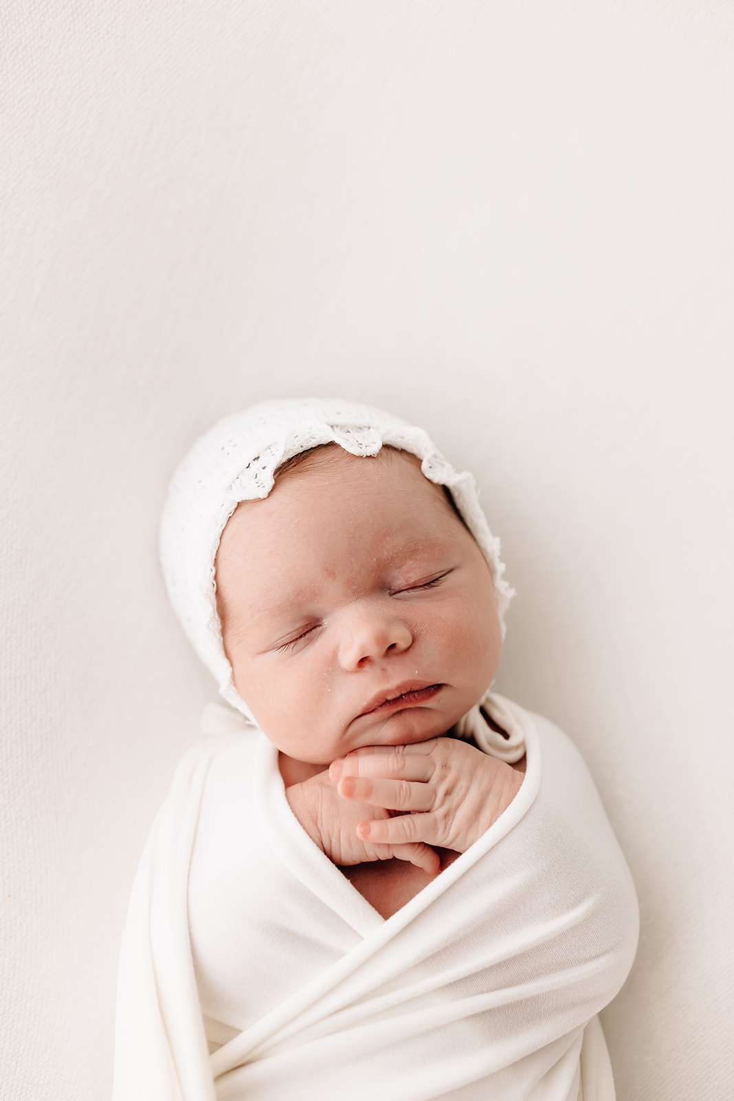 A newborn baby sleeps in a white swaddle and bonnet with hands crossed under her chin St Louis baby stores