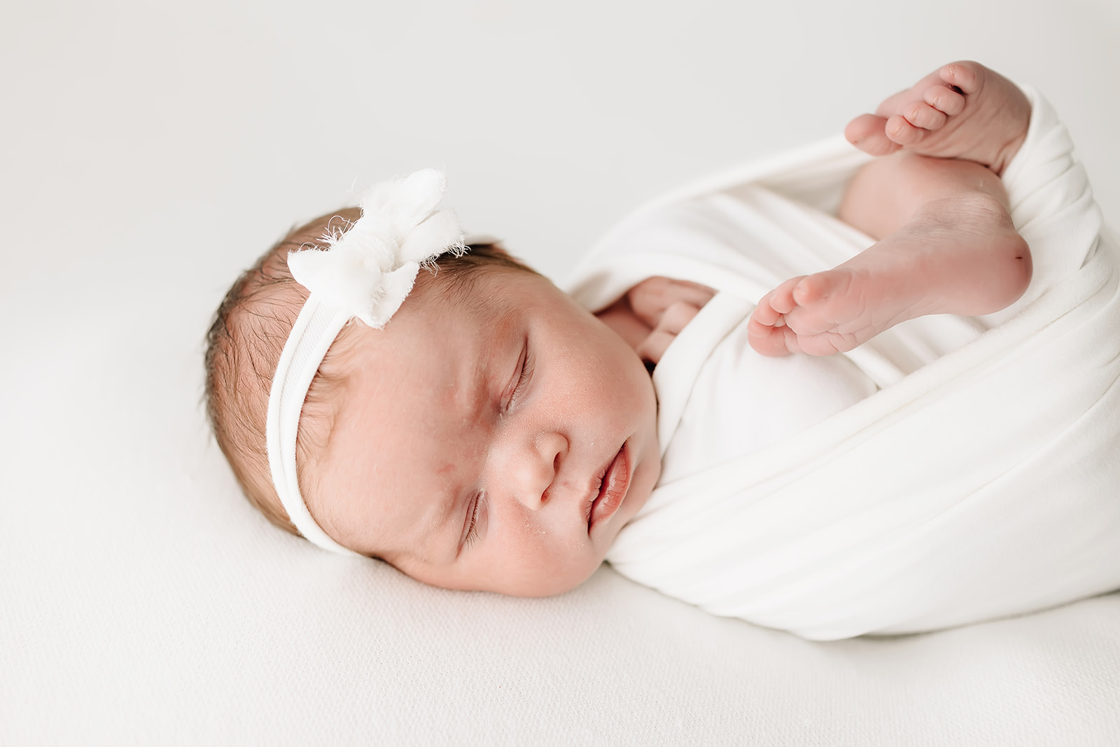 A newborn baby sleeps in a white swaddle with feet sticking out