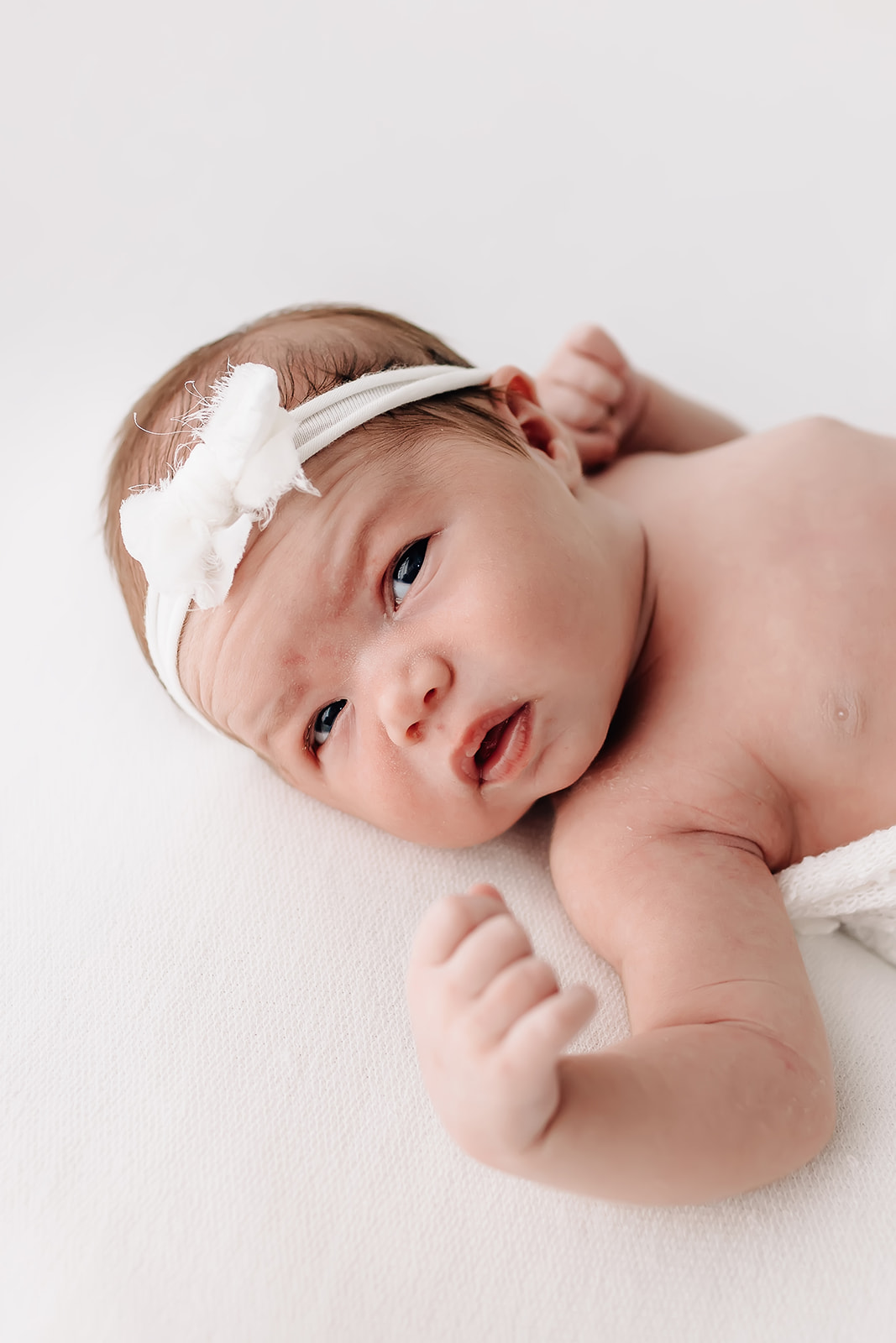 A newborn baby lays on a white bed with a white headband bow with eyes open