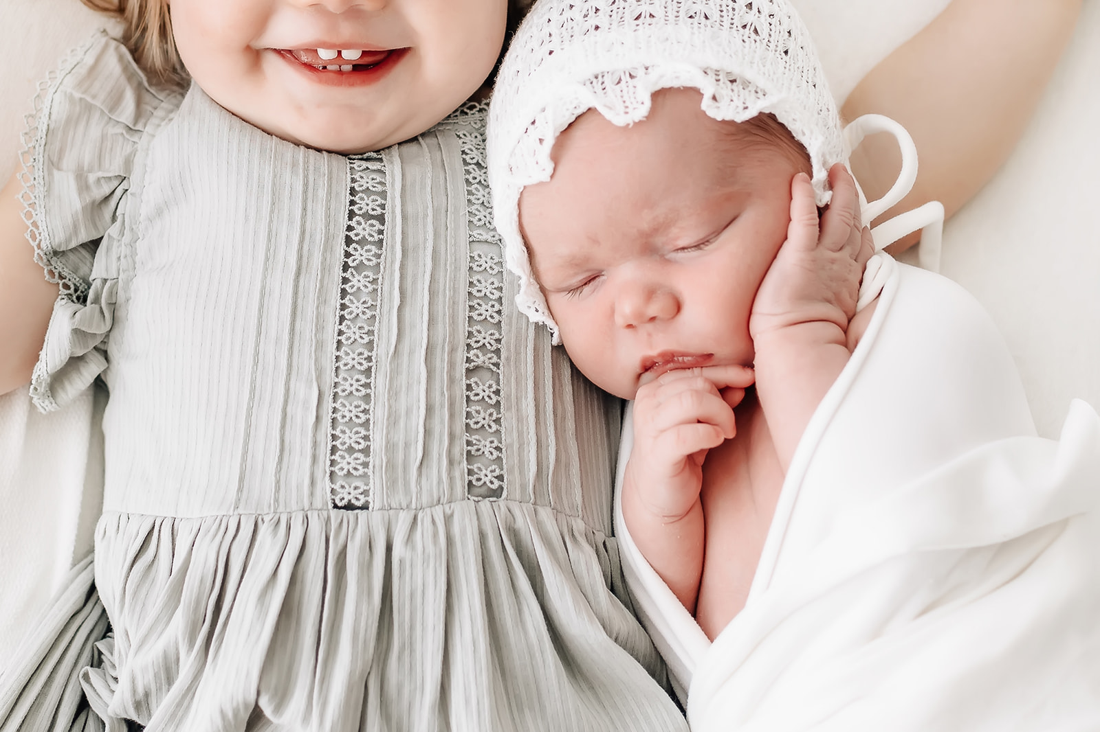A newborn baby in a white bonnet sleeps while cuddling against big sister