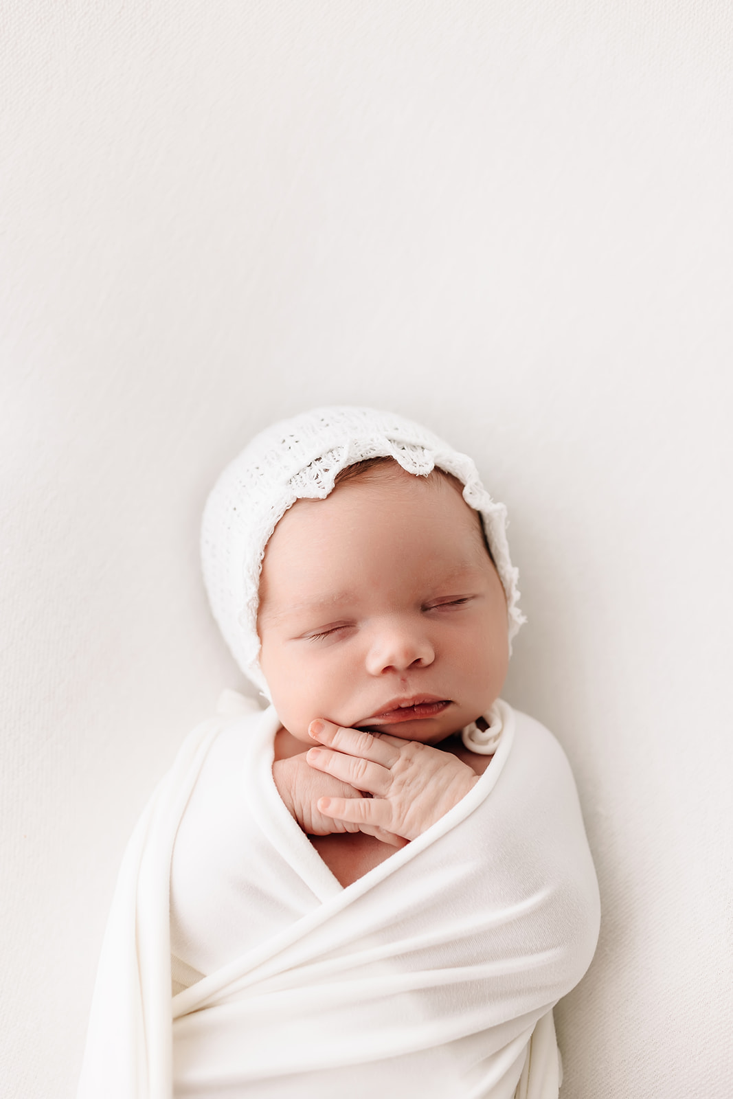 A newborn baby sleeps on its back with hands out in a white knit bonnet