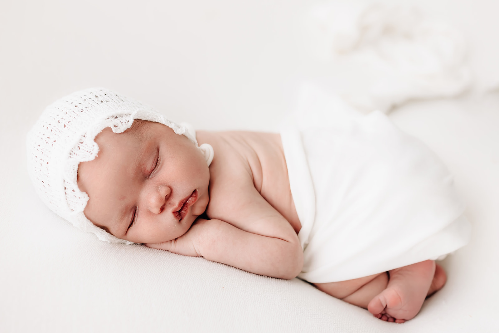 A newborn baby sleeps on a white bed wrapped in a blanket in a white bonnet St Louis Midwife