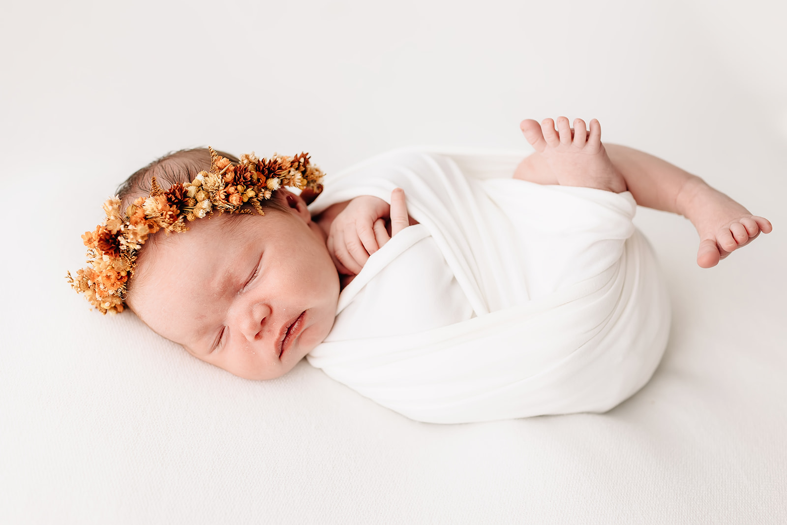A newborn baby wiggles out of a swaddle while sleeping in on a white bed with a floral headband