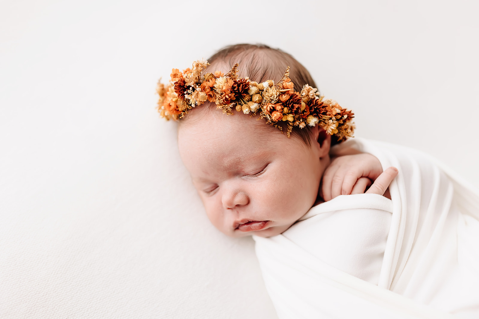 A newborn baby sleeps with fingers sticking out of a white swaddle with a floral headband daycare st louis