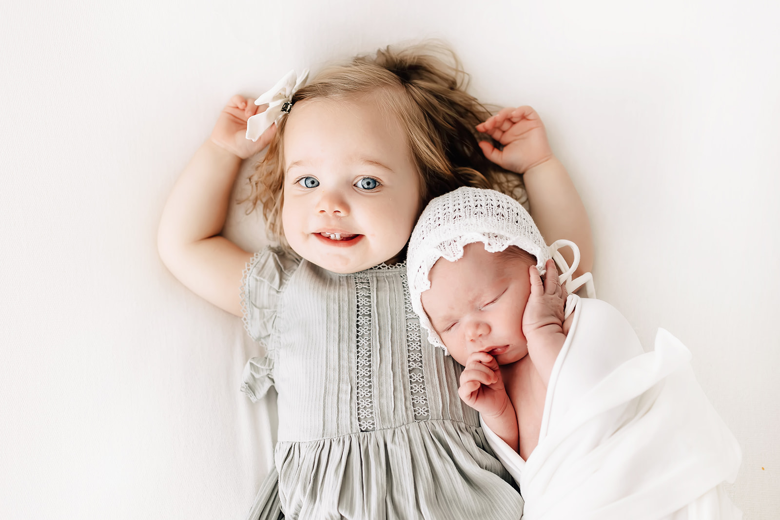 A young girl in a green dress lays on a bed while her newborn baby sibling sleeps against her lake st louis pediatricians