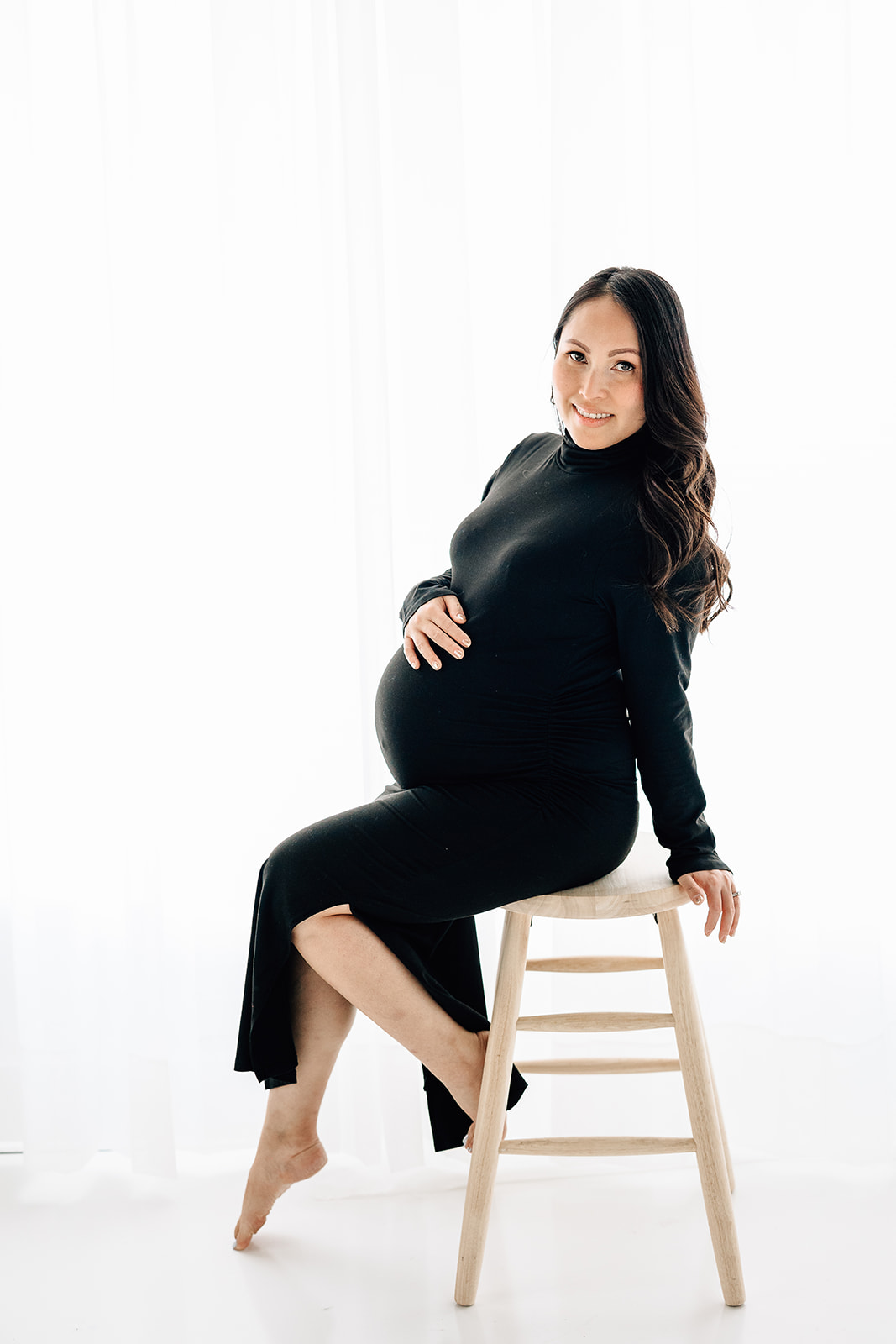A mom to be sits on a wooden stool in a studio holding her bump and looking over her shoulder