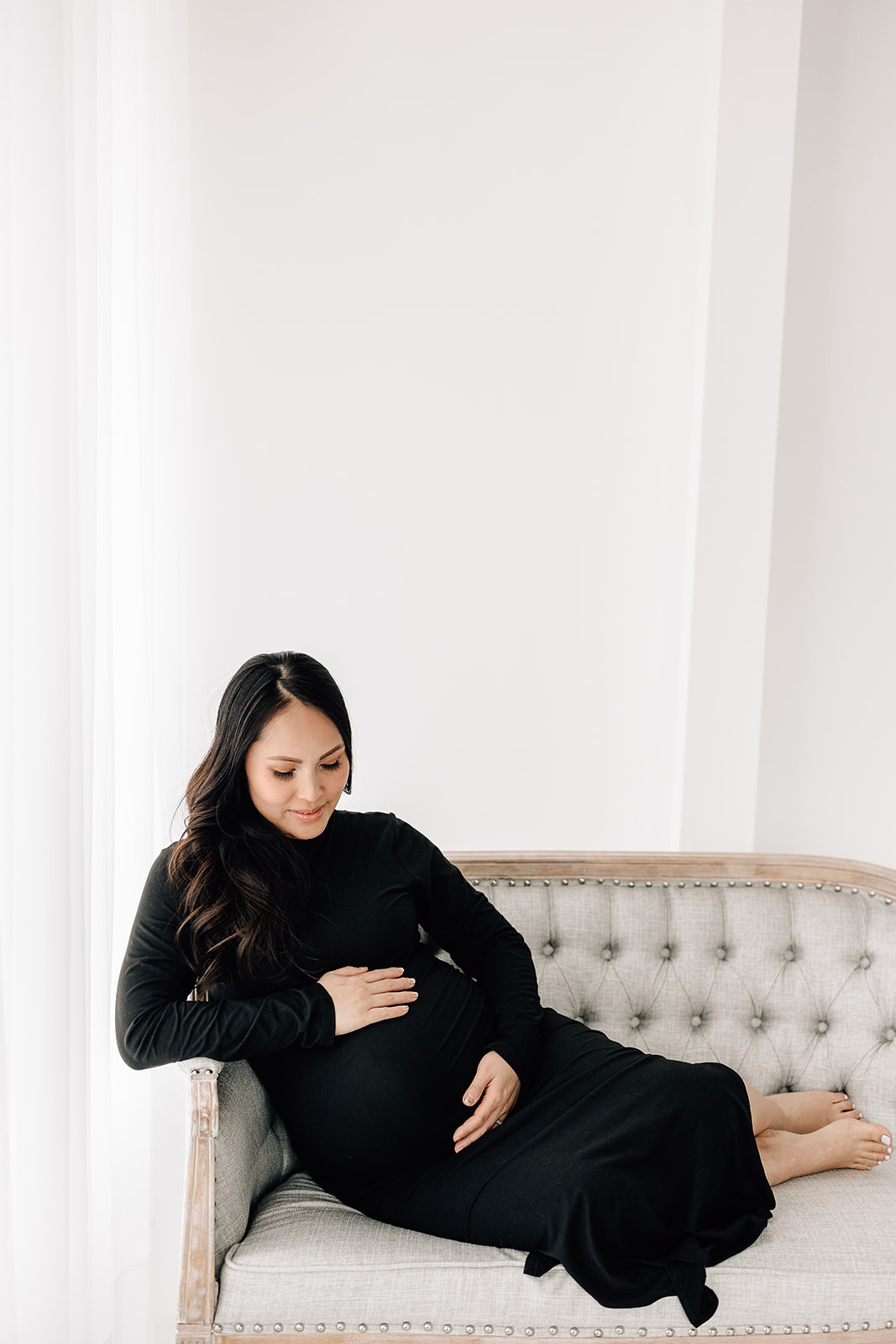 A mom to be sits across an antique bench in a studio holding her bump in a black maternity gown St. Louis Baby Shower Venues