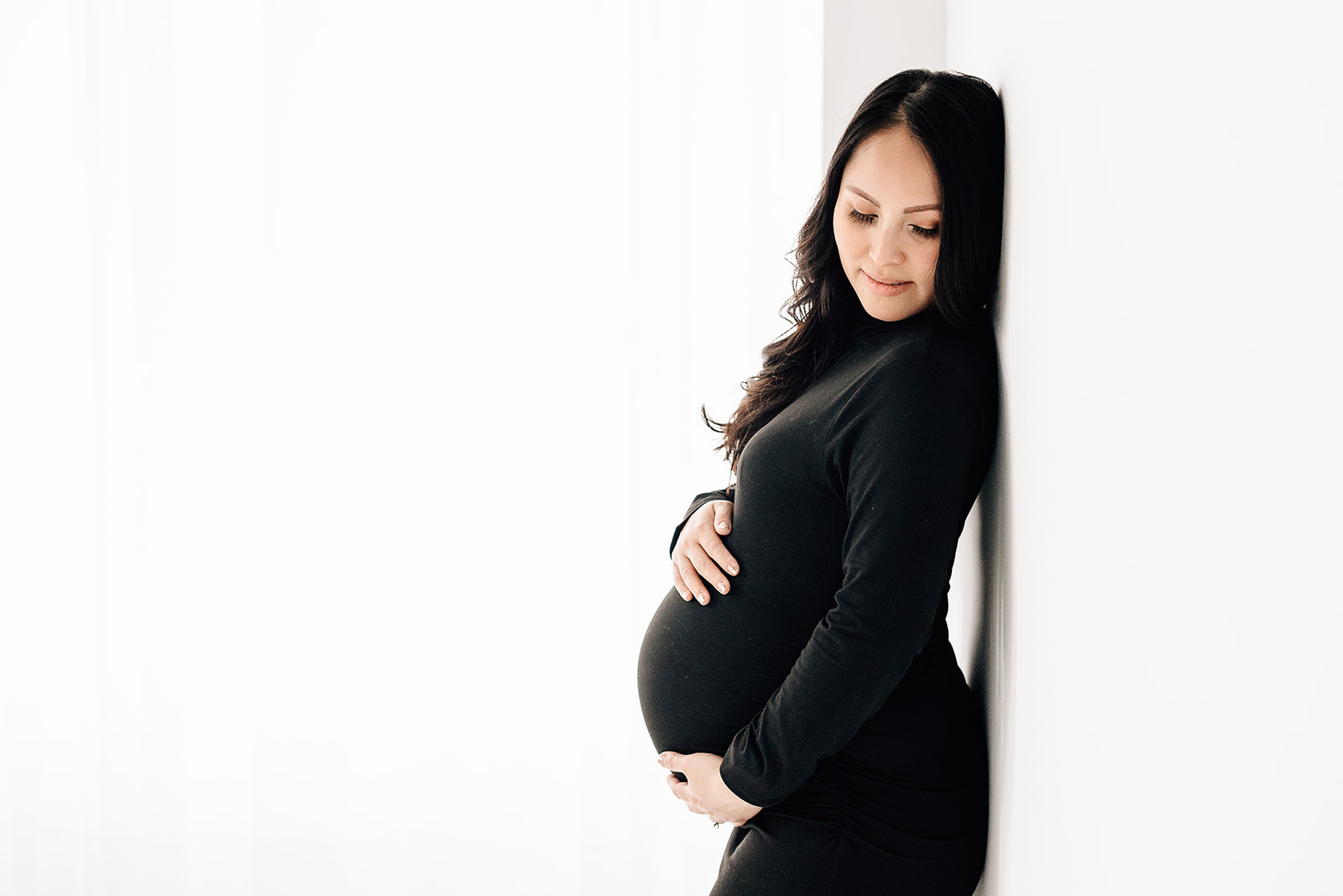 A mother to be in a black maternity gown leans against a wall in a studio St. Louis Baby Shower Venues