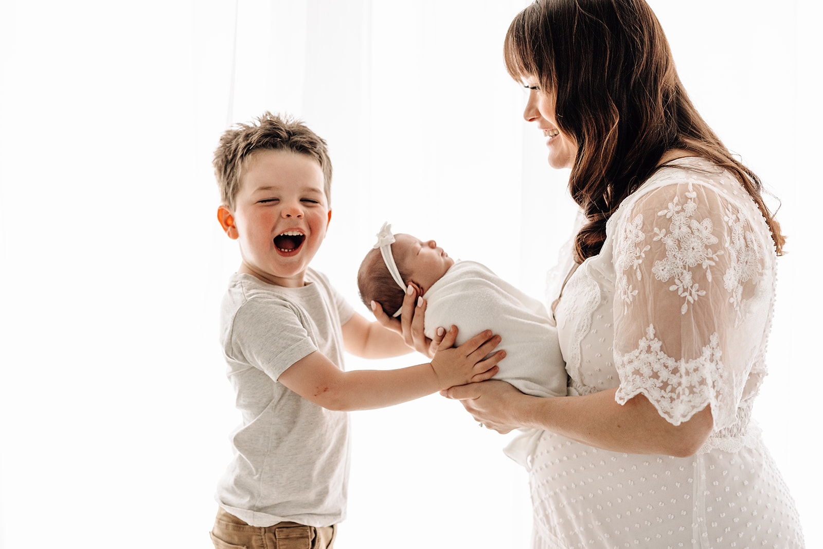 A young boy smiles as he places his hands on his newborn baby sister being held by mom in a studio St. Louis Nannies