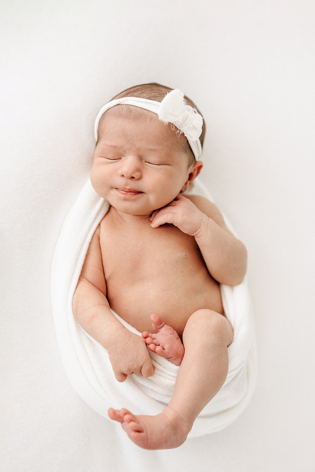 A newborn baby girl sleeps in a white blanket with a matching bow