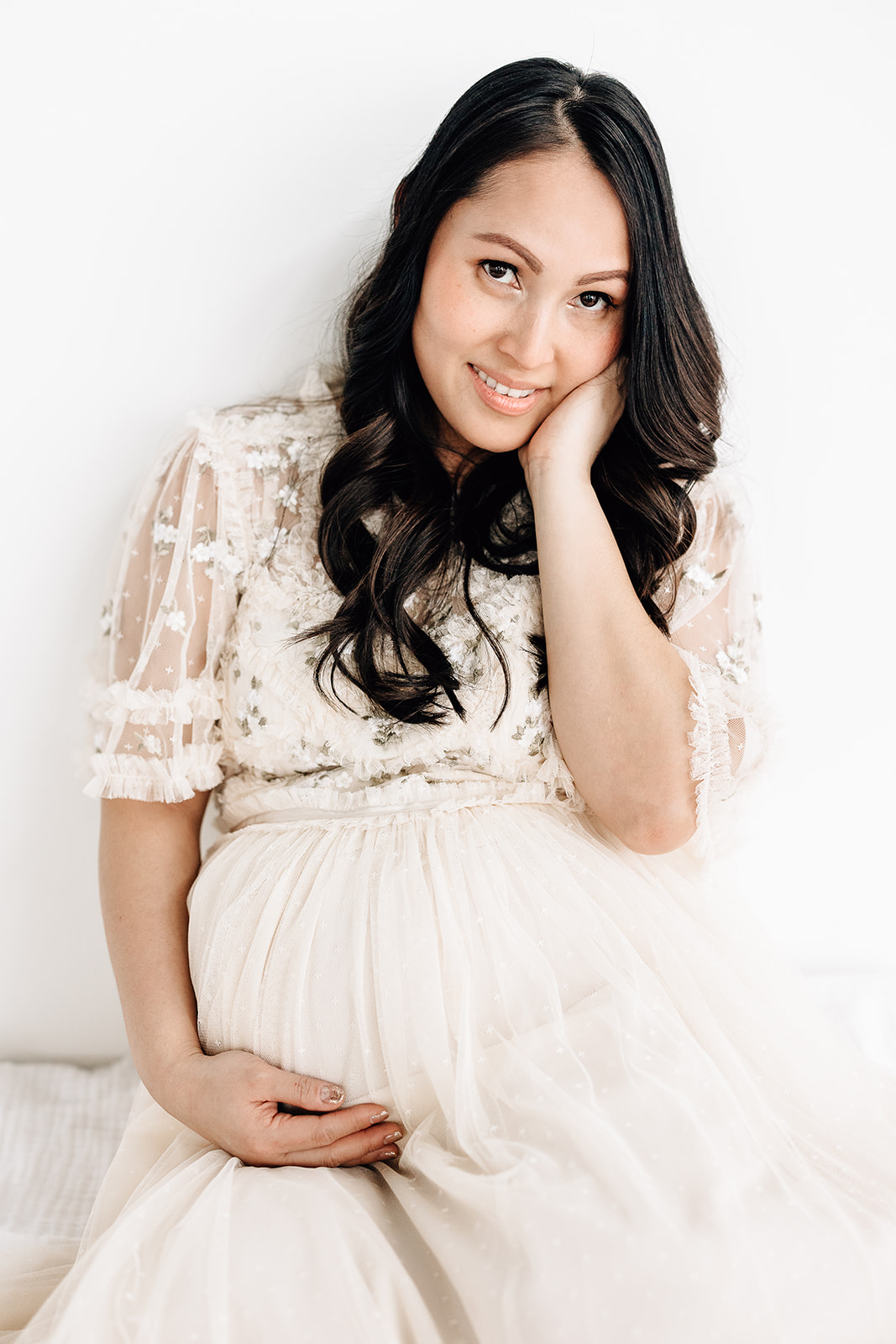 A mom to be sits against a wall with a hand under her bump in a white maternity dress Prenatal chiropractor St. Louis