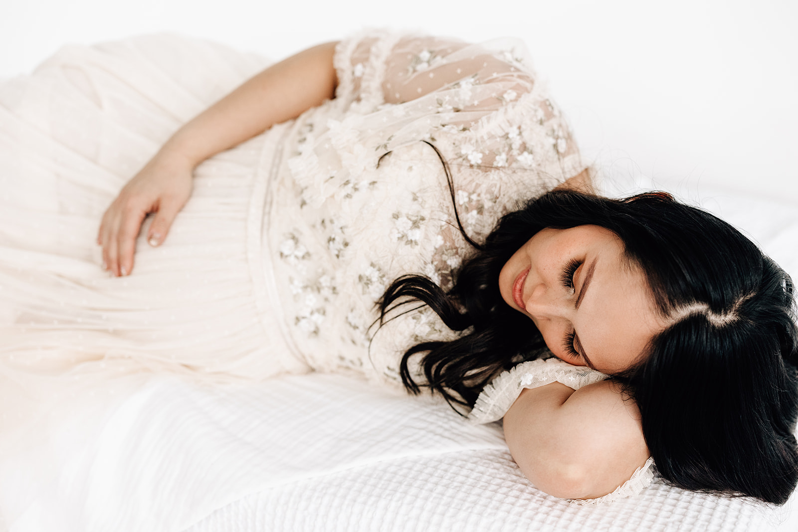 A mom to be in a white lace maternity gown lays across a bed in a studio Prenatal chiropractor St. Louis