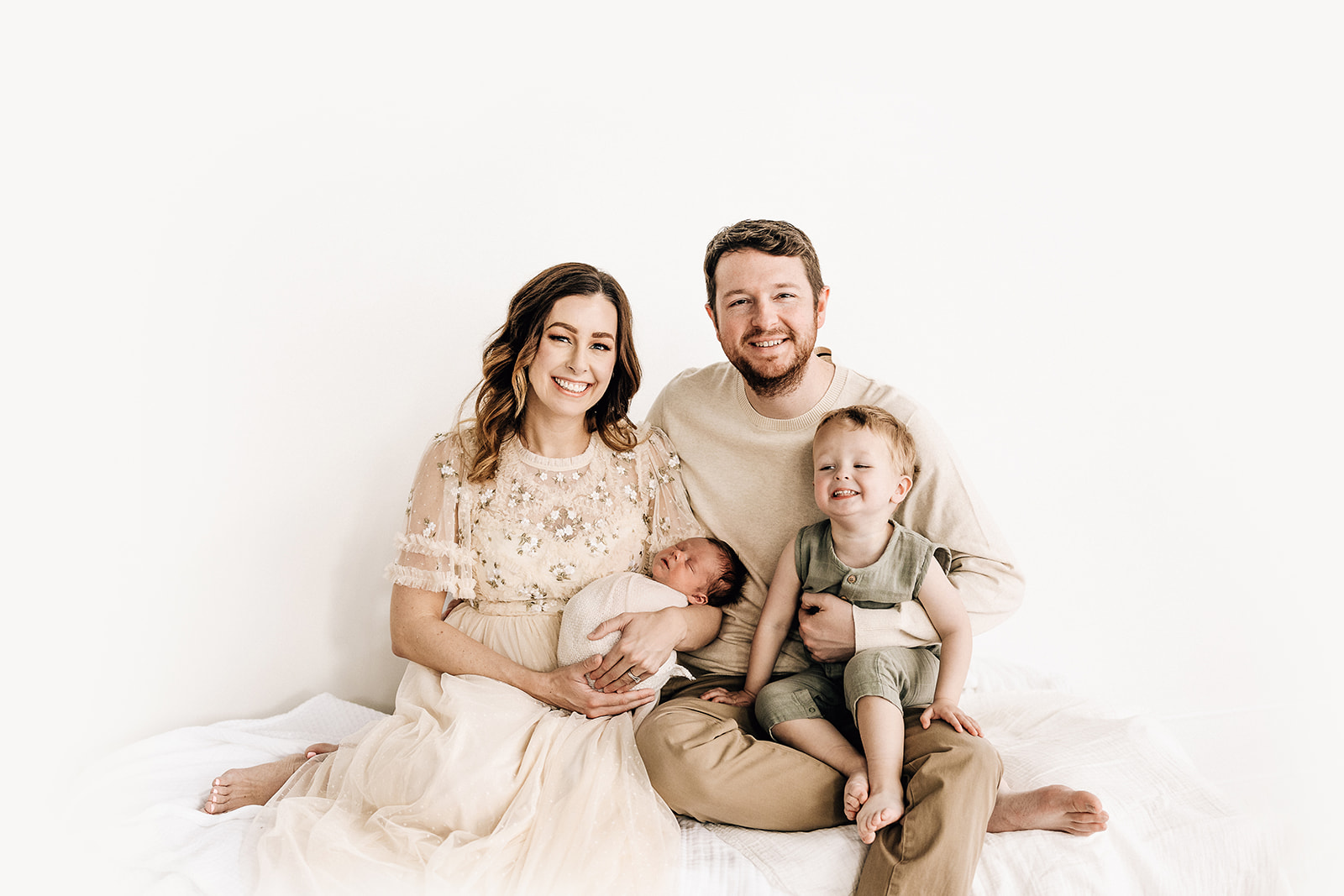 Mom and dad in beige outfits sit on a bed while mom holds their newborn baby child and their toddler son in a green onesie sits on dad's lap