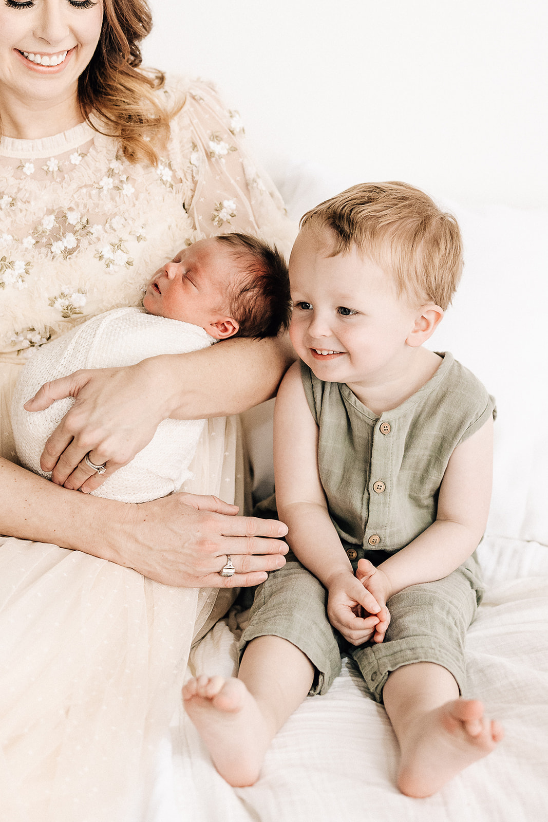 A toddler boy in a green onesie sits beside mom while she holds his sleeping newborn sibling St. Louis Babysitters