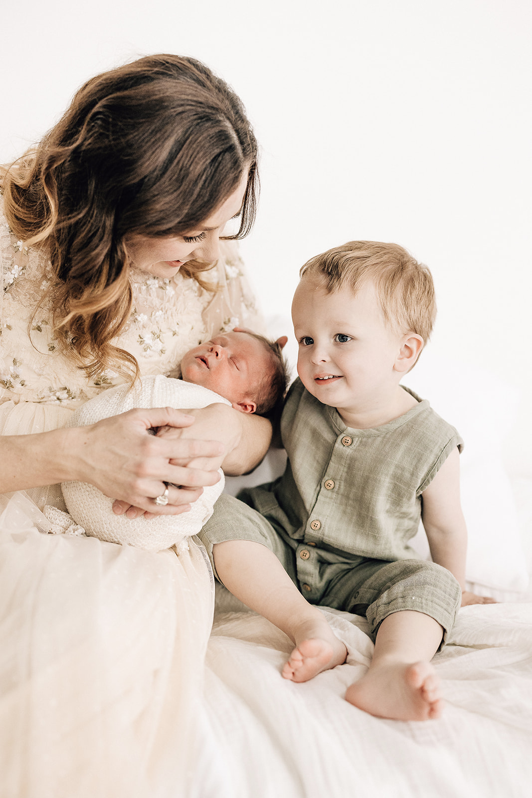 A mother sits on a bed with her toddler son while cradling her sleeping newborn St. Louis Babysitters