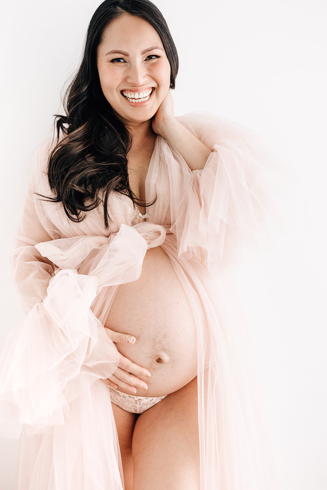 A mom to be laughs while standing in a studio wearing a pink maternity gown