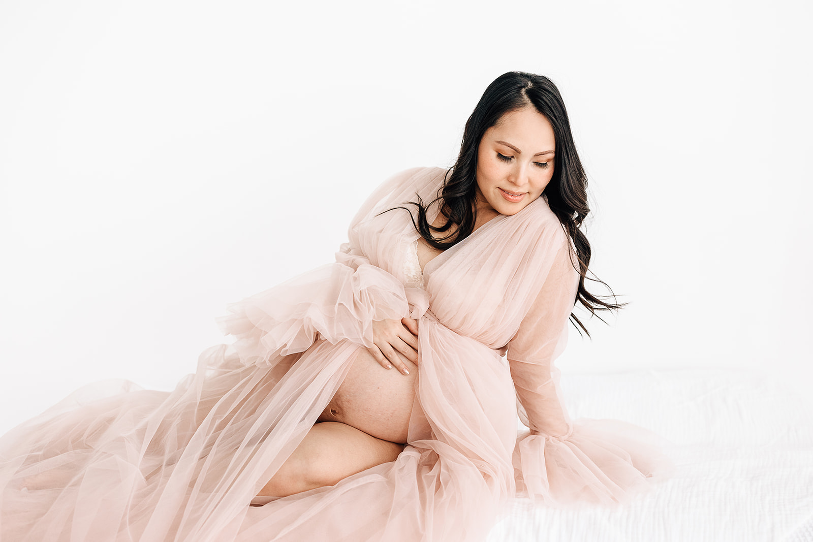 A mom to be lays across a bed in a pink maternity gown with bump exposed St. Louis Prenatal Massage