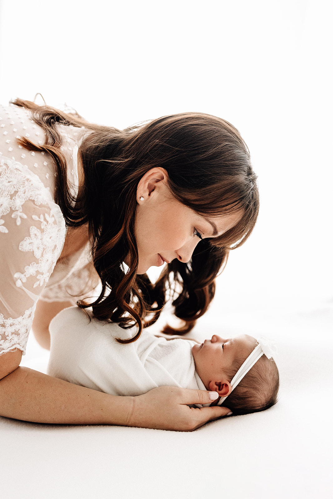 A new mom leans over her newborn baby on a white bed in a white lace bed Barefoot Midwife