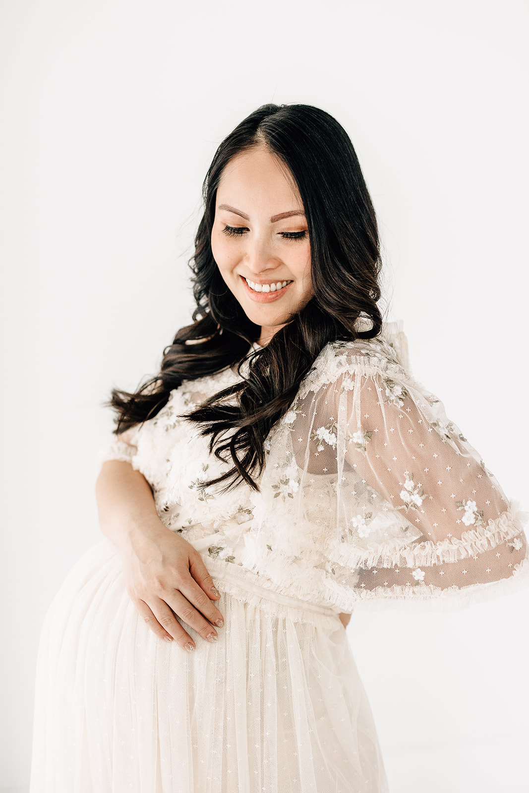A mom to be in a white lace maternity gown smiles down her shoulder while resting her arm on her bump
