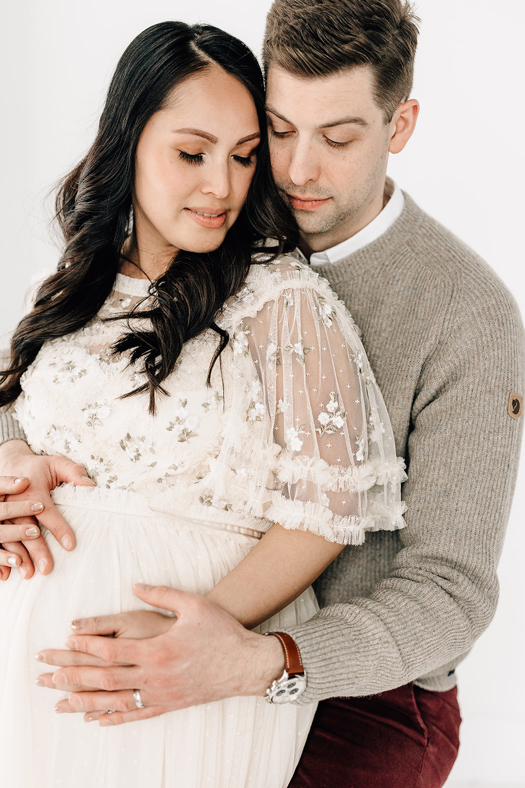 A mom to be leans into her partner while wearing a white lace maternity gown as dad wears a grey sweater and they rest their hands on the bump Mercy Birthing Center