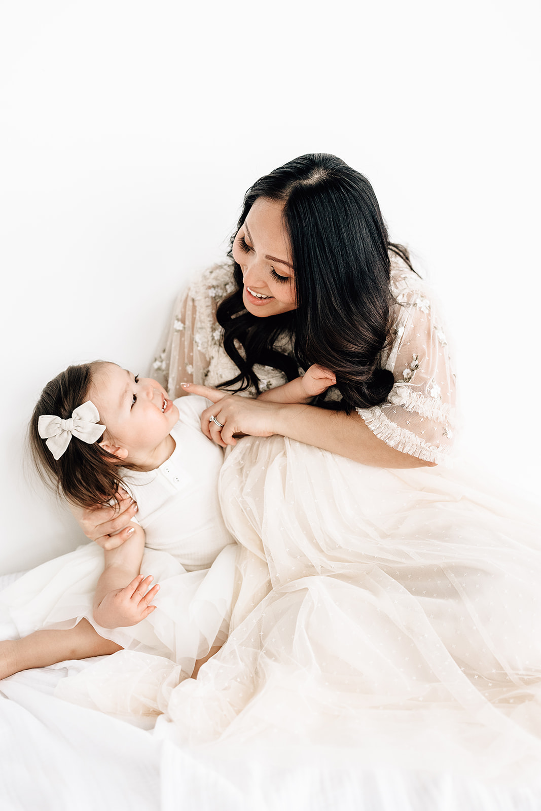 A pregnant mother in a lace maternity gown sits on a white bed playing with her toddler daughter in a white dress and hair bow before visiting Mercy Birthing Center