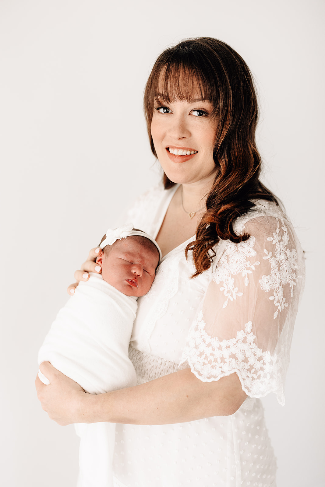 A mom in a white dress stands in a studio holding her sleeping newborn daughter in a white swaddle against her chest
