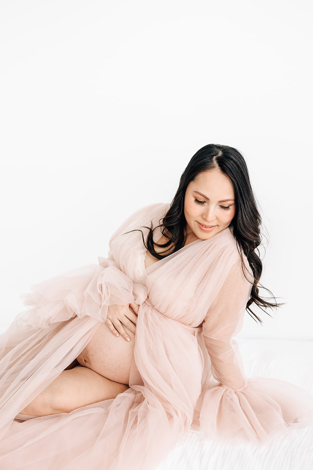 A mom to be with bump exposed in her pink velour maternity gown sits on a white bed in a studio Water Birth St. Louis