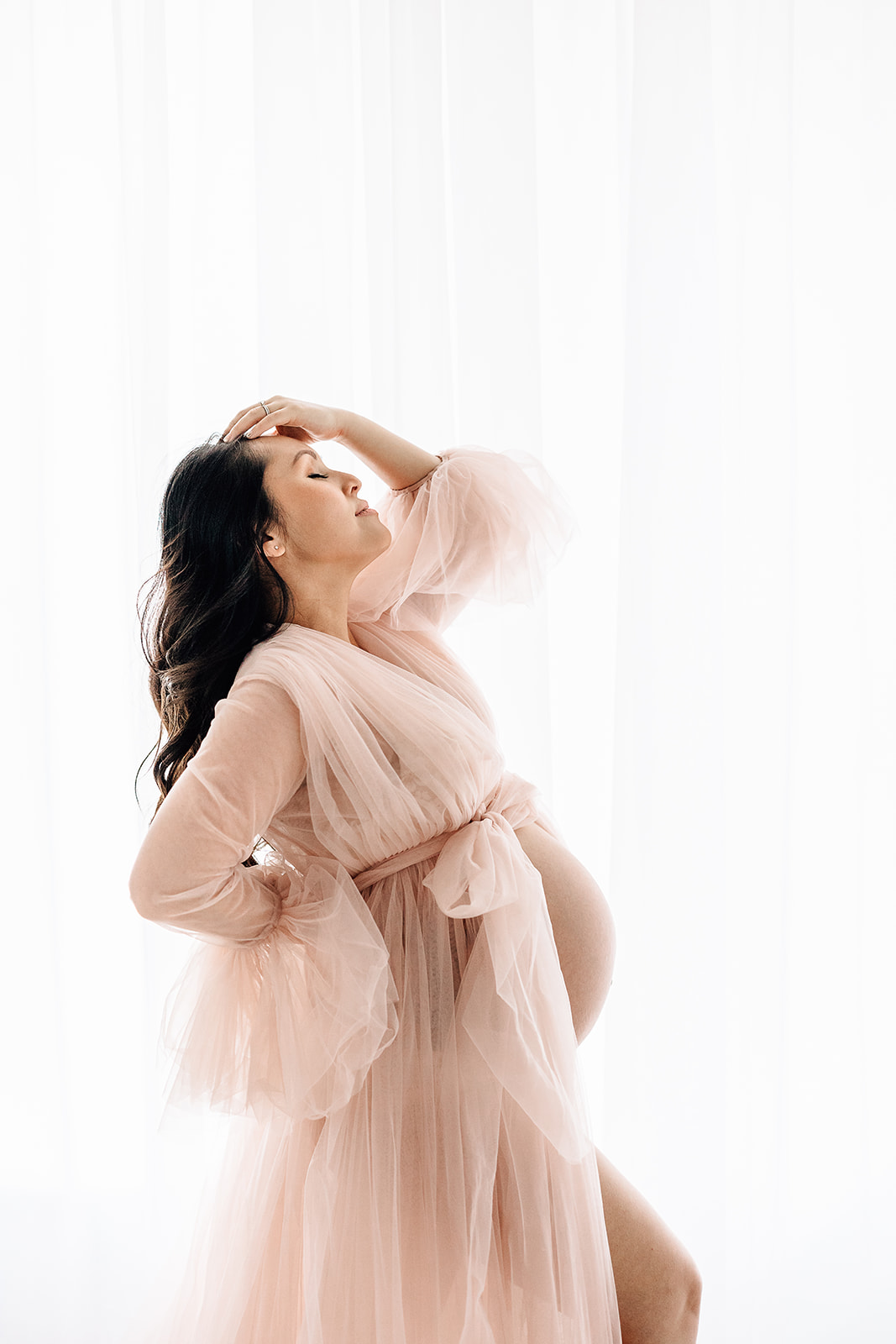 A mom to be stands in a window in a studio in a pink velour maternity gown with a hand on her head and another on her back before a Water Birth St. Louis