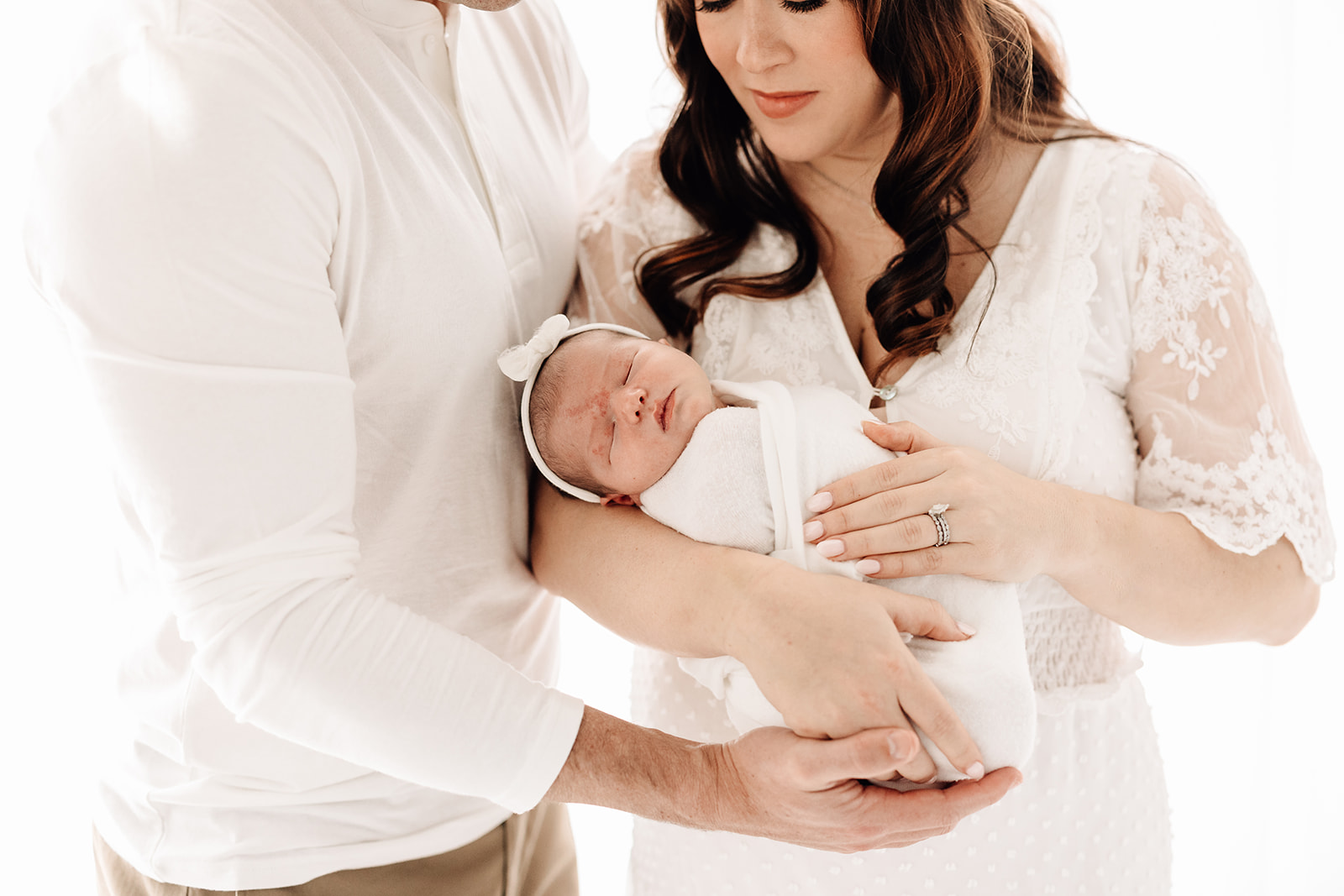 A mom in a lace dress cradles her sleeping newborn baby with dad while standing in a window