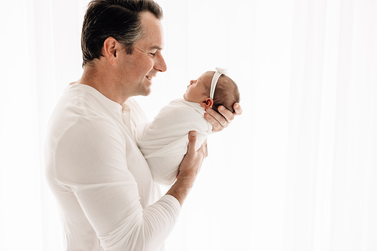 A new dad smiles down at his sleeping newborn daughter in his hands while standing in a window in a studio thanks to a Sleep Consultant St Louis