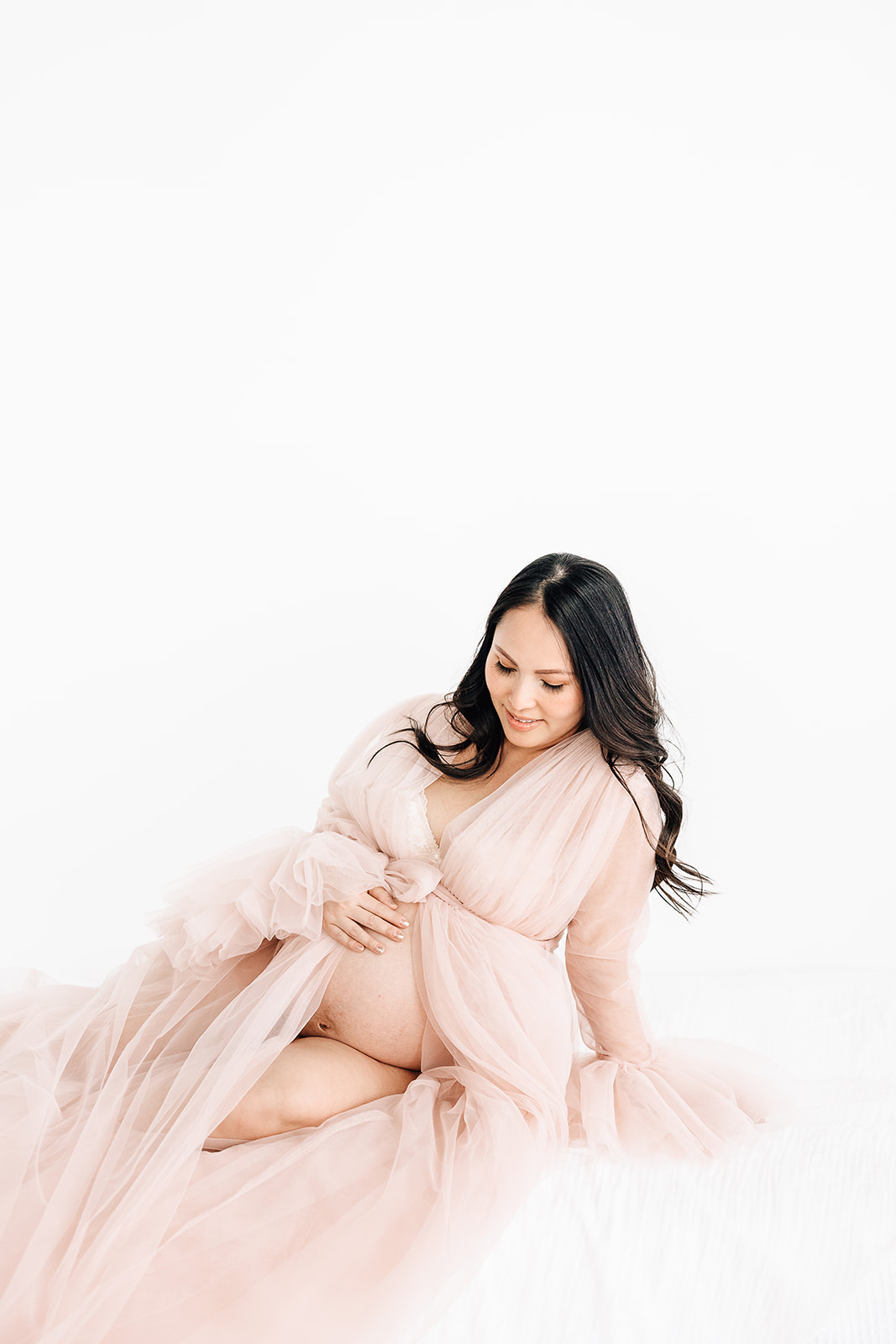 A mom to be in a pink maternity gown sits on a bed in a studio smiling down at her exposed bump after visiting a St. Louis OBGYN