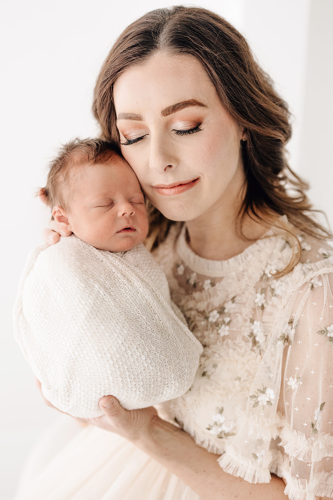 A mother in a beige dress stands in a studio nuzzling her sleeping newborn baby to her cheek