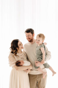 A mother and father in tan clothes stand in a studio window holding their toddler son and newborn baby after using 3D Ultrasound St. Louis