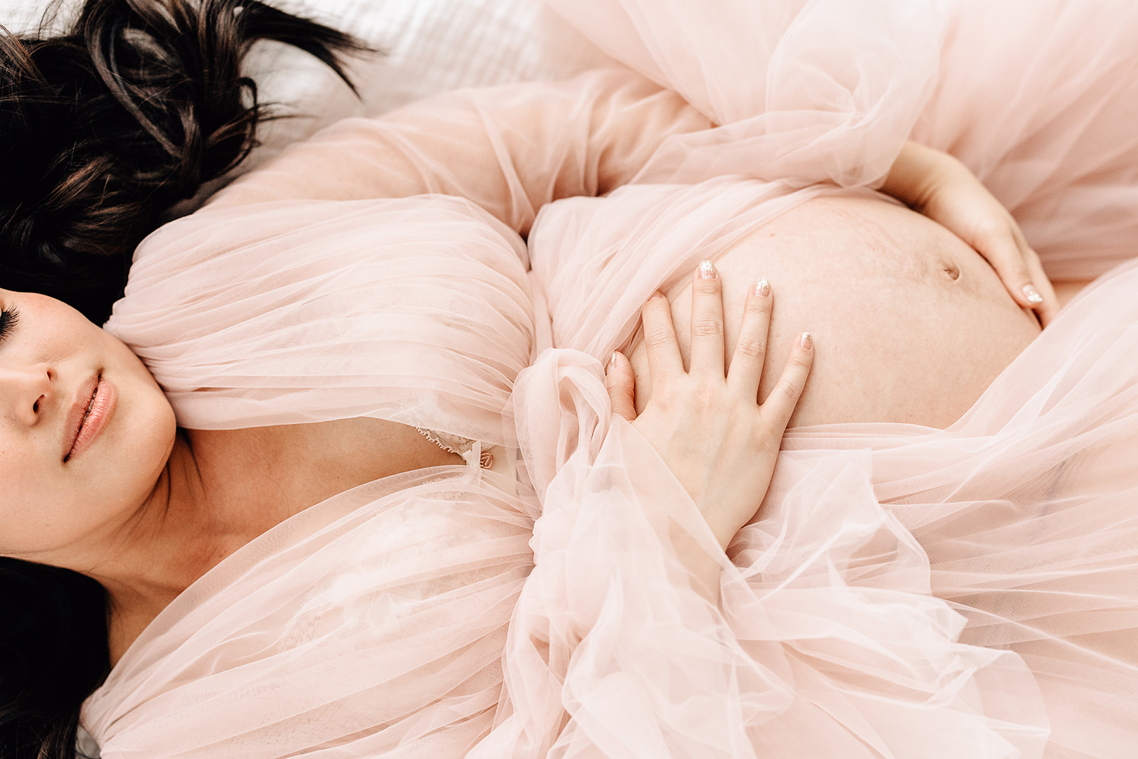 A mom to be lays on a bed in a pink velour maternity gown holding her bump thanks to a St. Louis Fertility Clinic