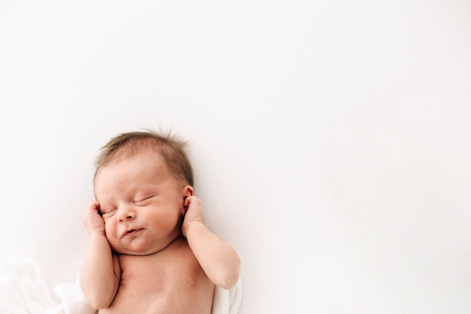 A newborn baby wiggles in its sleep on a white bed with hands up