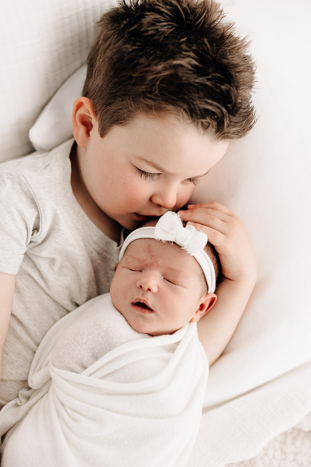 A young boy nuzzles his sleeping newborn baby sister while laying on a bed